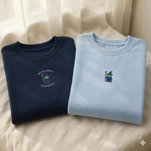 May include: A dark navy blue sweatshirt and a light blue sweatshirt, each featuring a small embroidered graphic of a green frog wearing a blue wizard hat and robe, holding a brown staff, with the text "OFF TO COMMIT SHENANIGANS" in white.