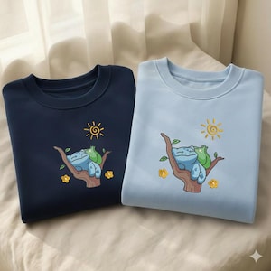 May include: Two crew neck sweatshirts, navy and light blue, with embroidered cartoon designs. Each features a character sleeping on a tree branch, with a sun and flowers. The sweatshirts are made of a soft material.