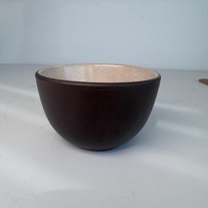 May include: A small, round bowl featuring a dark brown exterior with a smooth, matte finish. The interior of the bowl is a contrasting light beige colour with a speckled, textured appearance. The rim is thin and rounded, showing a clear transition between the dark outer surface and the lighter inner surface.