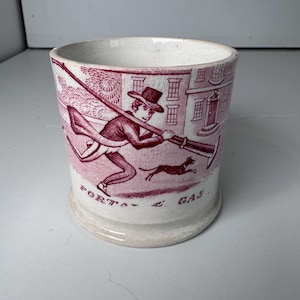 May include: A cream-colored ceramic mug featuring a maroon transferware print of a person in a top hat carrying a long pole over their shoulder, accompanied by a small dog. The text "PORTABLE GAS" is printed in maroon at the base of the design.