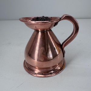 May include: A polished copper vessel with a wide, flared rim and a single curved handle attached to the side. The body of the container is rounded and tapers toward the top, featuring a metallic finish with small dark spots across the surface.