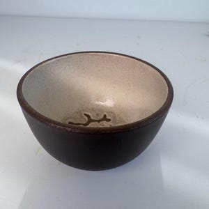 May include: A small, round ceramic bowl with a dark brown exterior and a speckled, light beige interior. The bottom of the bowl features a dark brown, branch-like or twig-shaped graphic design centred inside the basin.