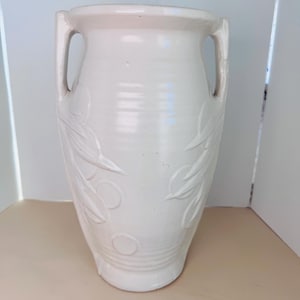 May include: A tall, off-white ceramic vase featuring two integrated side handles and a relief pattern of leaves and circular shapes embossed on the exterior. The vessel has a wide, flared rim and a smooth, glossy finish over the entire surface.