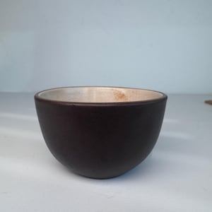May include: A small, rounded ceramic bowl featuring a dark brown, matte exterior finish and a contrasting light cream-coloured interior. The bowl has a simple, curved silhouette with a smooth rim and a deep, open basin for holding various items.