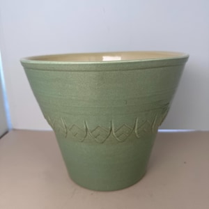 Può includere: Un vaso da fiori in ceramica color verde salvia con un bordo largo e svasato e una base affusolata. L'esterno presenta una fascia orizzontale con motivi in rilievo a forma di chevron che circondano la sezione centrale. L'interno è color crema.
