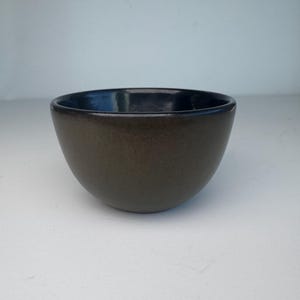 May include: A small, round ceramic bowl with a smooth, dark brown exterior and a glossy, deep black interior. The bowl features a simple, curved silhouette with a wide opening and a solid base, showcasing a consistent, dark, earthy colour palette.