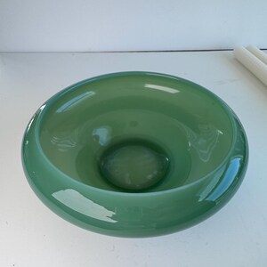 Peut inclure: Un bol large et peu profond en verre vert sauge translucide et lisse. Le bol présente un bord épais et arrondi ainsi qu'un centre concave profond avec une finition polie qui reflète la lumière sur sa surface circulaire incurvée.