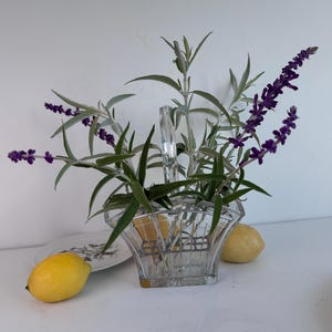 May include: A clear glass basket-style vase with a central handle and a metal grid insert, containing sprigs of purple salvia flowers with green leaves. Two yellow lemons are positioned on either side of the glass vase on a white surface.