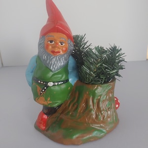Könnte beinhalten: Eine Gartenzwerg-Figur mit roter Mütze, blauen Ärmeln und grüner Tunika mit schwarzem Gürtel, die neben einem braunen Baumstumpf-Pflanzgefäß steht. Das Gefäß enthält künstliche grüne Tannennadeln und einen kleinen roten Pilz.