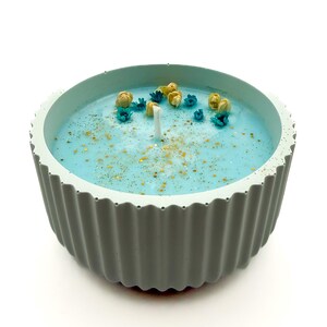 Peut inclure: Une bougie ronde bleu clair dans un récipient en béton gris cannelé. La bougie a une cire bleu clair avec des paillettes dorées et de petites fleurs décoratives bleues et jaunes. Une mèche blanche est centrée.