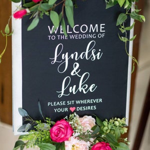 May include: A black chalkboard sign with white text that reads "Welcome to the wedding of Lyndsi & Luke. Please sit wherever your heart desires." The sign is decorated with pink and white flowers.