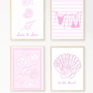 May include: Four framed art prints with pink line drawings on white and pink backgrounds. The prints feature seashells, a bikini on a clothesline, and the text "Sun & Sea" and "to the Beach". The frames are a light wood color.