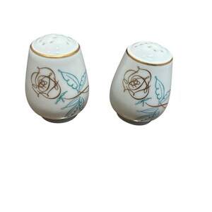 May include: Two white ceramic salt and pepper shakers with a gold rim and a floral design in gold and teal. The shakers have a rounded shape and a series of holes on top for dispensing.