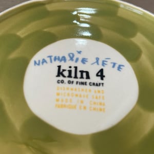 May include: A close-up of a green ceramic bowl with a white circular label in the center. The label features the text "Nathalie Lete kiln 4 CO. OF FINE CRAFT" in blue and black. Additional text indicates the bowl is dishwasher and microwave safe, made in China.