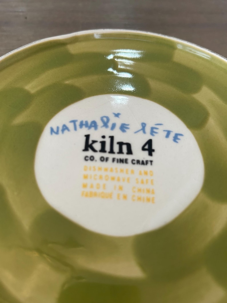 May include: A close-up of a green ceramic bowl with a white circular label in the center. The label features the text "Nathalie Lete kiln 4 CO. OF FINE CRAFT" in blue and black. Additional text indicates the bowl is dishwasher and microwave safe, made in China.