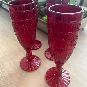 Puede incluir: Cuatro copas de vino de color rojo rubí con detalles en relieve. Las copas tienen una forma alta y delgada y una base redonda. Las copas están dispuestas sobre una superficie de madera de color claro, con un fondo borroso de vegetación.