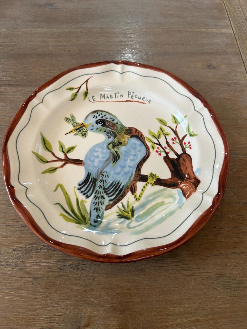 May include: A decorative ceramic plate with a bird design. The plate features a blue bird perched on a brown branch with green leaves and red berries. The rim is brown, and the plate has the text "LE MARTIN P&Ecirc;CHEUR".