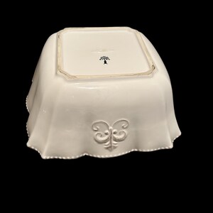 Op de afbeelding: Een witte, vierkante keramische serveerschaal met een geschulpte rand en een decoratief reli&euml;f. De kom heeft een gestileerd bloemenembleem aan de onderkant en een decoratief reli&euml;f aan de zijkant.