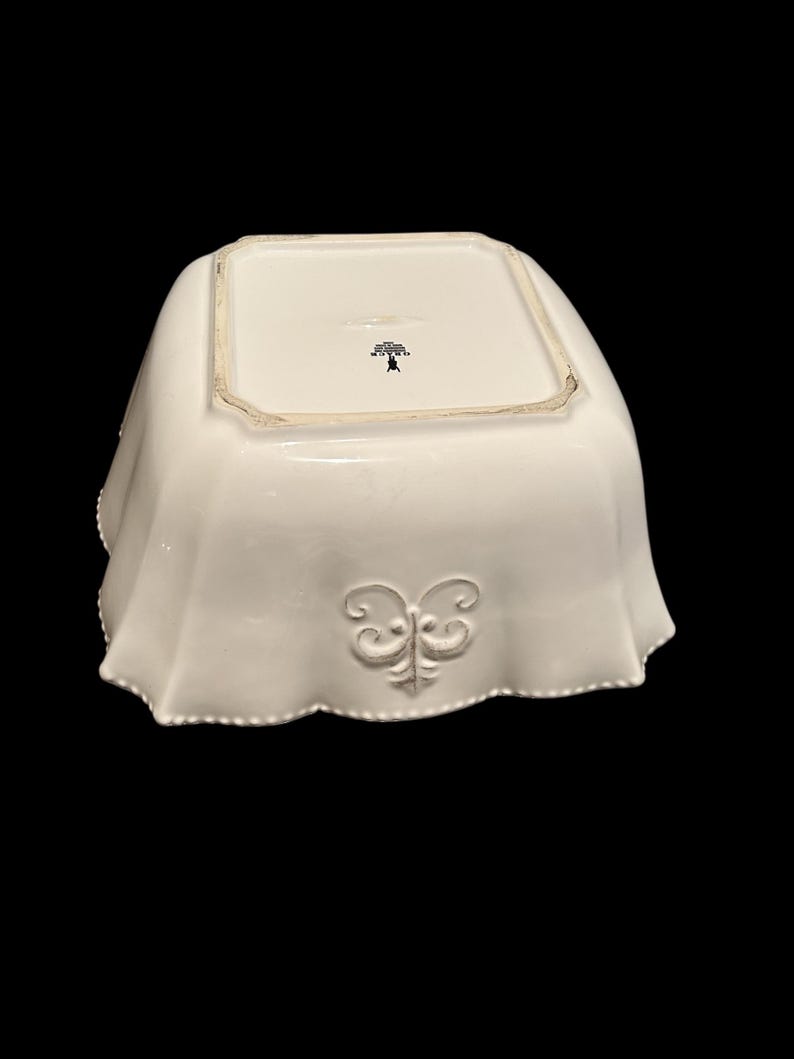 Op de afbeelding: Een witte, vierkante keramische serveerschaal met een geschulpte rand en een decoratief reli&euml;f. De kom heeft een gestileerd bloemenembleem aan de onderkant en een decoratief reli&euml;f aan de zijkant.