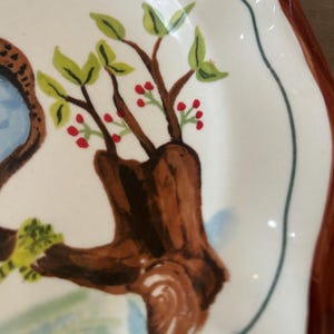 May include: A white ceramic plate with a brown rim and a scalloped edge. The plate features a hand-painted design of a tree trunk with green leaves and red berries. A blue and brown design is visible on the left side.