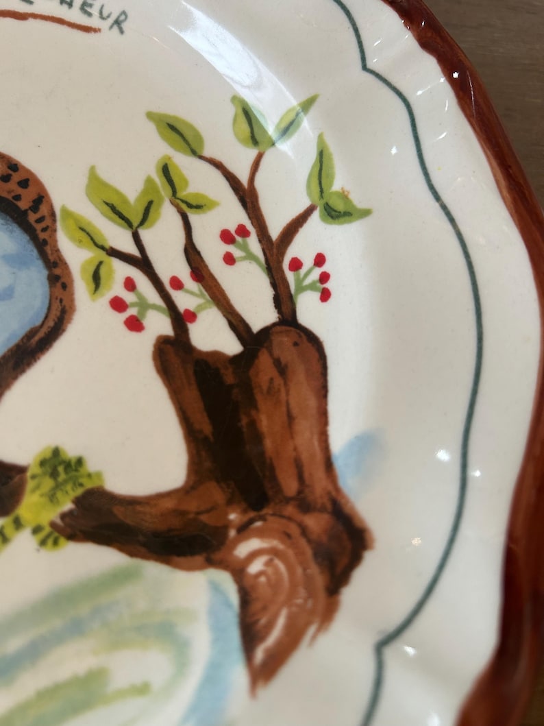 May include: A white ceramic plate with a brown rim and a scalloped edge. The plate features a hand-painted design of a tree trunk with green leaves and red berries. A blue and brown design is visible on the left side.