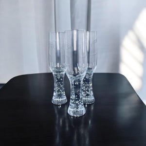 May include: Four clear glass beer glasses with bubble-like designs in the stem. The glasses are on a dark wooden table with a white curtain in the background. The glasses are empty.