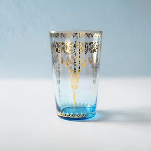 May include: A clear glass with a blue base and gold filigree design. The gold pattern is ornate and symmetrical, with a repeating design around the rim and a central motif. The glass is likely for drinking.