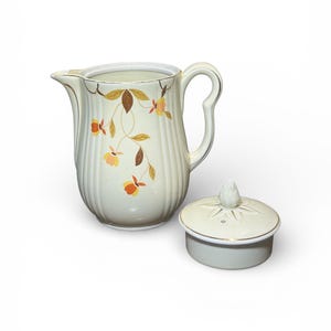 May include: Cream-colored ceramic pitcher with a matching lid. The pitcher features a floral design with orange and yellow flowers and brown leaves. The lid has a gold trim and a decorative knob.