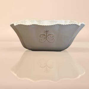 Op de afbeelding: Een grijze keramische kom met een geschulpte rand en een decoratief reli&euml;fontwerp. De kom heeft een brede, open vorm en een glad, reflecterend oppervlak. Het reli&euml;fontwerp is gecentreerd op de voorkant van de kom.