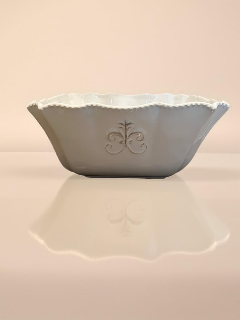 Op de afbeelding: Een grijze keramische kom met een geschulpte rand en een decoratief reli&euml;fontwerp. De kom heeft een brede, open vorm en een glad, reflecterend oppervlak. Het reli&euml;fontwerp is gecentreerd op de voorkant van de kom.