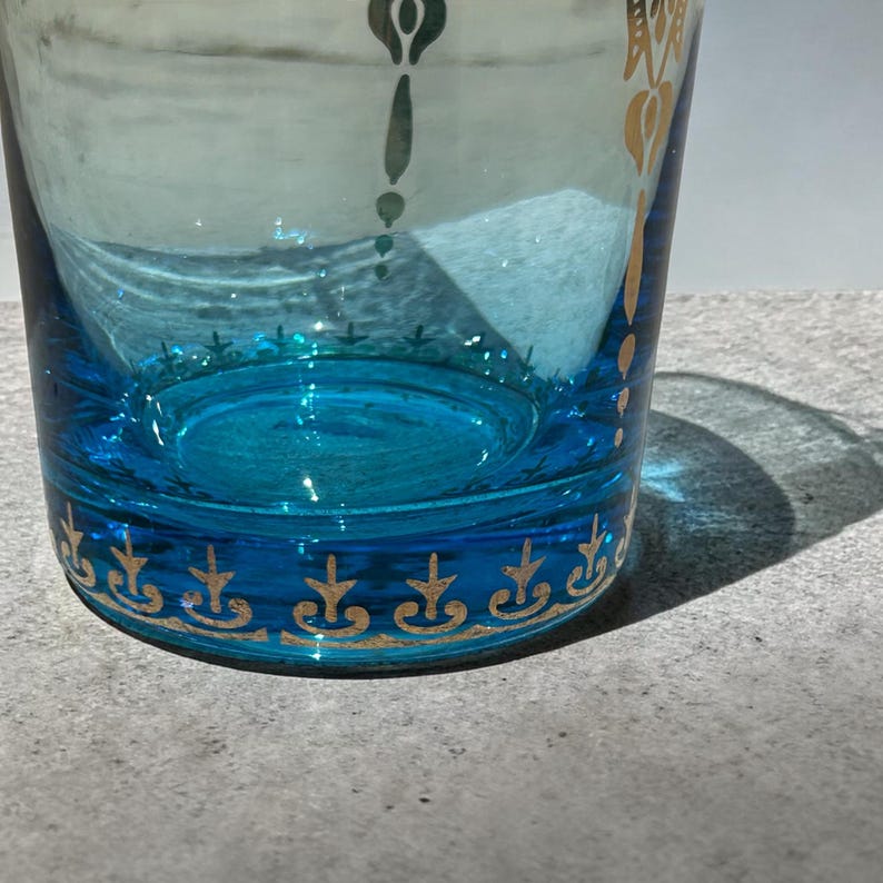 May include: A clear glass with a turquoise base and accents. The glass features gold decorative patterns around the base and upper rim. The glass is partially filled, and the design is visible against a light background.