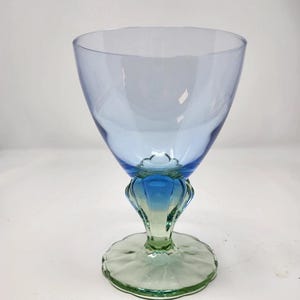 May include: A tall, elegant wine glass with a blue bowl and a decorative stem. The stem transitions from blue to green, resting on a scalloped base. The glass is clear, allowing light to pass through.