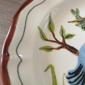 May include: Cream-colored ceramic plate with a scalloped edge and a brown rim. The plate features a hand-painted design of a blue bird, green leaves, and brown branches. A thin dark blue line borders the inner edge.