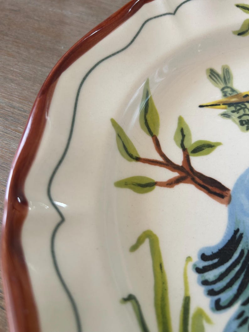 May include: Cream-colored ceramic plate with a scalloped edge and a brown rim. The plate features a hand-painted design of a blue bird, green leaves, and brown branches. A thin dark blue line borders the inner edge.