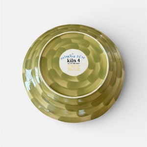 May include: A ceramic plate with a green and tan painted design. The plate's underside is visible, showcasing a circular pattern of brushstrokes. The center features a white label with the text "Nathalie Lete kiln 4 CO. OF FINE CRAFT".