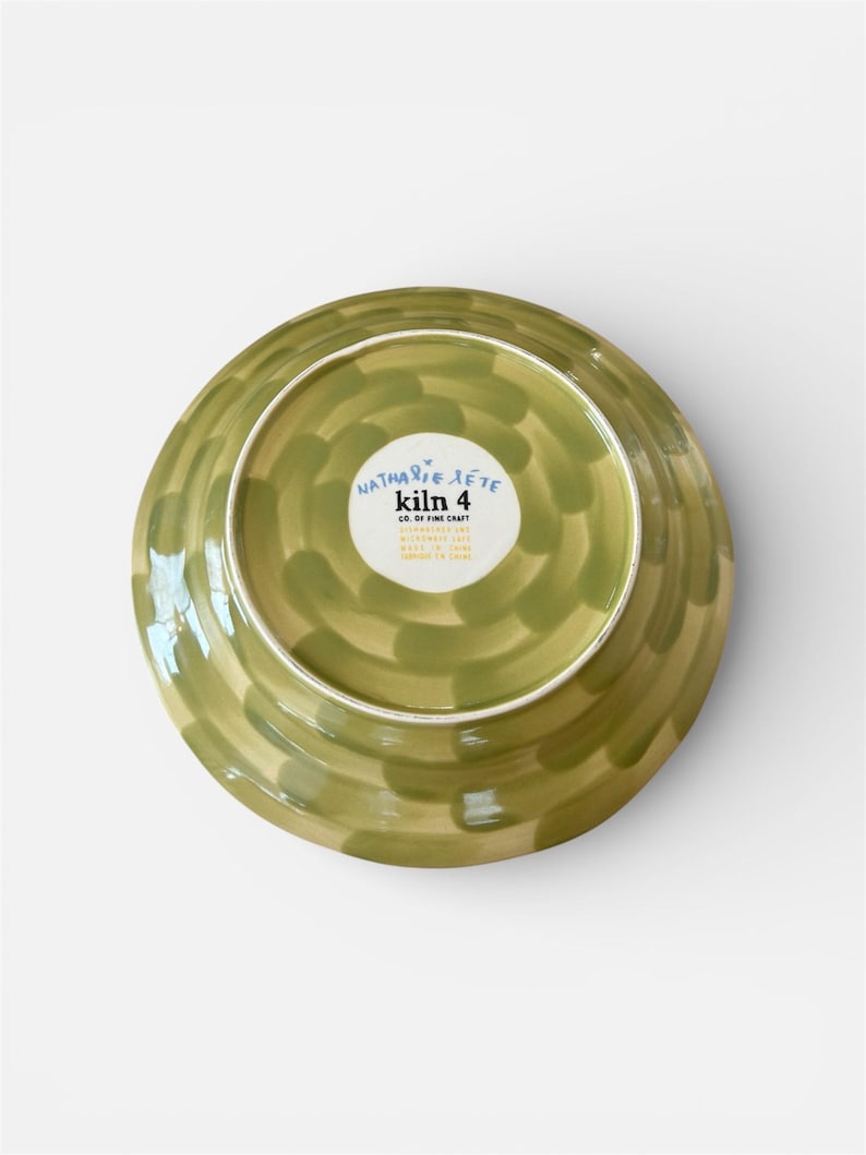 May include: A ceramic plate with a green and tan painted design. The plate's underside is visible, showcasing a circular pattern of brushstrokes. The center features a white label with the text "Nathalie Lete kiln 4 CO. OF FINE CRAFT".