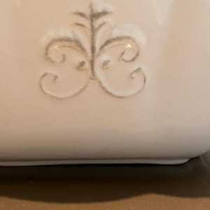 Op de afbeelding: Close-up van een witte keramische container met een decoratief, in reli&euml;f aangebracht bloemmotief in een gedempte grijze kleur. De bovenrand heeft een geschulpte rand. De container staat op een neutrale achtergrond.
