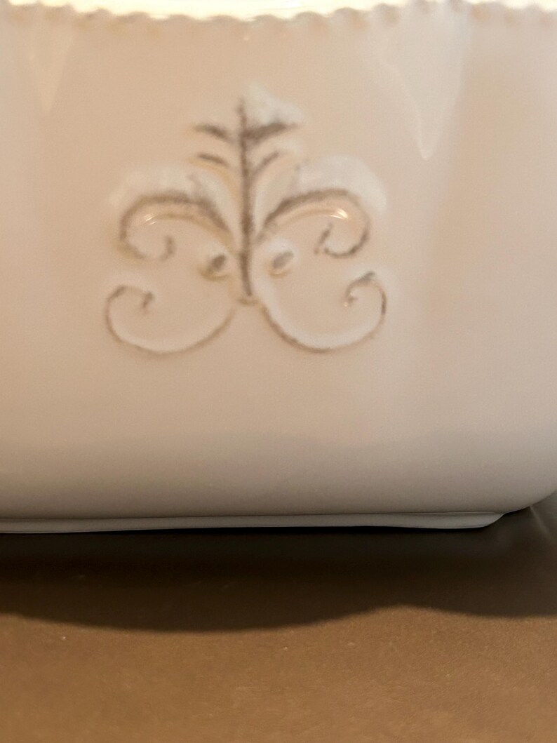 Op de afbeelding: Close-up van een witte keramische container met een decoratief, in reli&euml;f aangebracht bloemmotief in een gedempte grijze kleur. De bovenrand heeft een geschulpte rand. De container staat op een neutrale achtergrond.