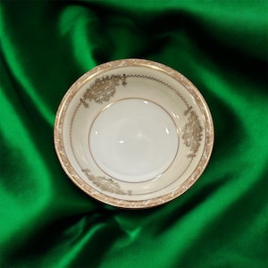 May include: A small, round, off-white ceramic bowl with a gold-colored floral and scroll design around the rim. The bowl has a smooth, glossy interior and is set against a green background.