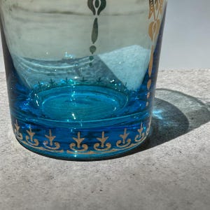 May include: A clear glass with a turquoise base and accents. The glass features gold decorative patterns around the base and upper rim. The glass is partially filled, and the light reflects off the surface.