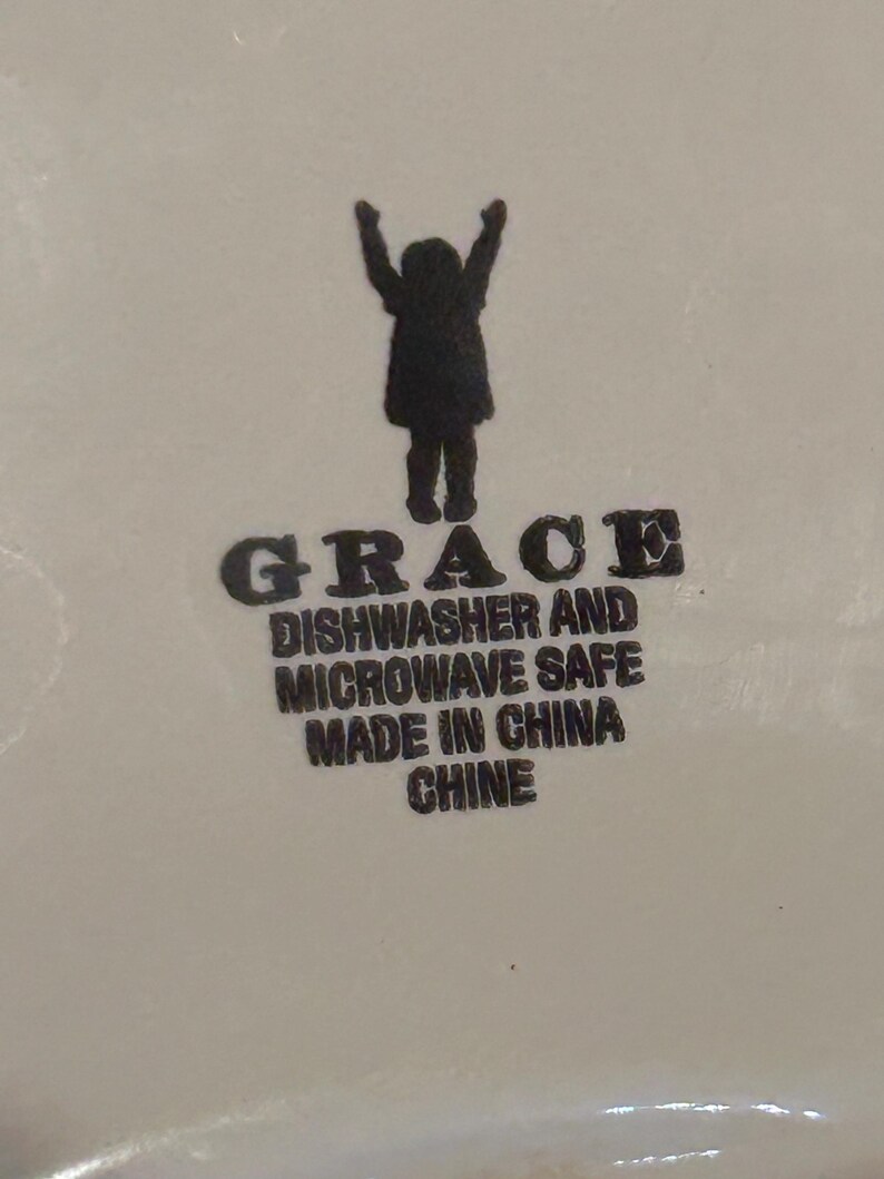 Op de afbeelding: Een wit keramisch oppervlak met het woord "GRACE" in vette, zwarte letters. Onder het woord "GRACE" staan de woorden "DISHWASHER AND MICROWAVE SAFE MADE IN CHINA". Een silhouet van een persoon met opgeheven armen staat boven het woord "GRACE".