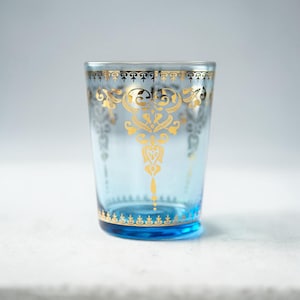 May include: A light blue glass with gold filigree designs. The glass features ornate gold patterns around the rim, a central design, and a base border. The glass is approximately 4 inches tall.