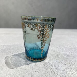 May include: A teal-colored glass with ornate gold filigree designs. The glass has a flared shape and a dark teal base with gold accents. The intricate patterns add a touch of elegance to this decorative glassware.