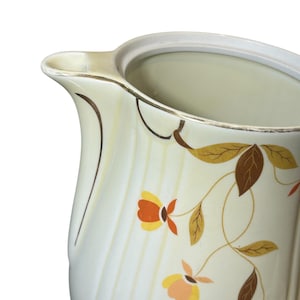 Puede incluir: Jarra de cerámica color crema con un diseño floral de flores y hojas naranjas, amarillas y marrones. La jarra tiene un pico curvo y un borde blanco, con líneas verticales y detalles marrones.