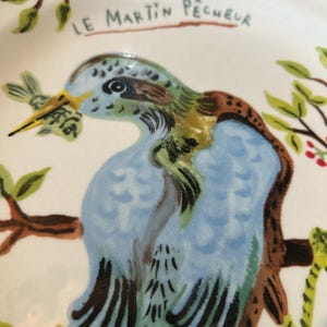 May include: A white ceramic plate with a hand-painted illustration of a blue, green, and brown bird perched on a branch. The bird is holding a small fish in its beak. The text "LE MARTIN P&Ecirc;CHEUR" is above the bird.