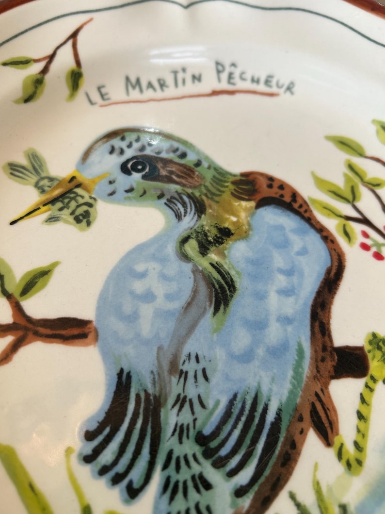May include: A white ceramic plate with a hand-painted illustration of a blue, green, and brown bird perched on a branch. The bird is holding a small fish in its beak. The text "LE MARTIN P&Ecirc;CHEUR" is above the bird.