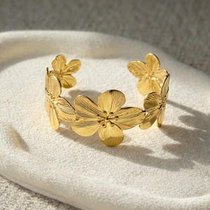 May include: A gold-colored cuff bracelet featuring a floral design. The bracelet is open and wraps around the wrist, with multiple detailed flower shapes. The flowers have textured petals and are arranged in a continuous pattern around the band.