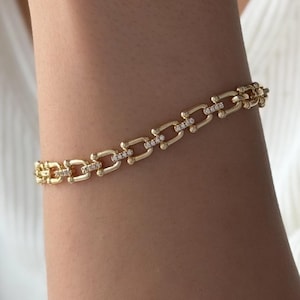 May include: A gold chain bracelet featuring rectangular links with rounded edges, where every other link is accented with a row of small, clear, sparkling stones set along the center, creating a repeating pattern of metal and stone.