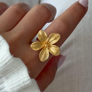 以下が含まれることがあります： 5枚の花びらを持つ大きな花の形をしたゴールドトーンの指輪。花びらには中心から放射状に広がる線状のテクスチャーが施されており、中心部には小さな隆起した球状のディテールがあります。指輪は指にはめられ、花のモチーフが強調されています。