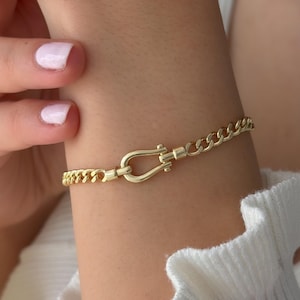 May include: A gold-toned chain link bracelet featuring a central horseshoe-shaped clasp. The bracelet consists of uniform interlocking metal links that connect to the U-shaped closure, creating a continuous loop around the wrist area.